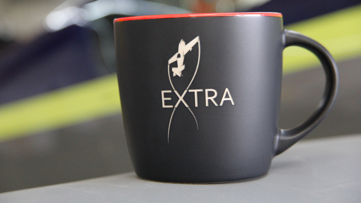 Produktbild der Extra Aircraft Keramiktasse. Außenfarbe: Mattschwarz, innenfarbe: Rocket-Red (glänzend). Die Tasse ist außen beidseitig mit dem offiziellen Extra Aircraft Logo graviert. Der Hintergrund ist unscharf, was die Tasse in den Mittelpunkt rückt.